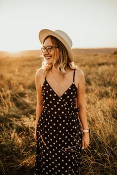 A fair-skinned blonde girl in a dress with polka dots in a hat walks through the meadow among the tall grass in the summer at sunset.