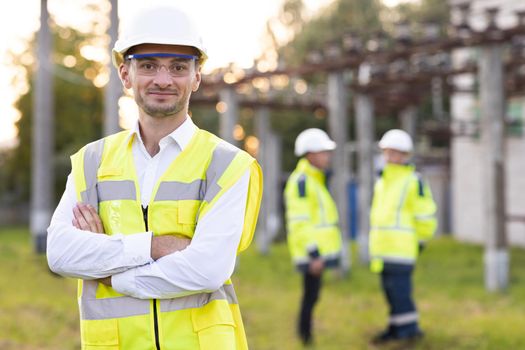 Portrait of male engineer technologist standing among high voltage electrical lines with her arms crossed over her chest. Man in protective helmet and uniform smiling and looking at camera.