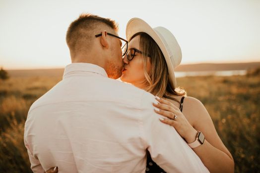 A man with a woman in a hat hug and kiss in the tall grass in the meadow. A couple of fair-haired fair-skinned people in love are resting in nature in a field at sunset.