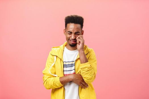 Portrait Lifestyle. Studio shot of silly sad african american timid guy in yellow trendy jacket crying heart out whiping tears from eyes and whining feeling lonely after being rejecting by ex-girlfriend over pink wall.