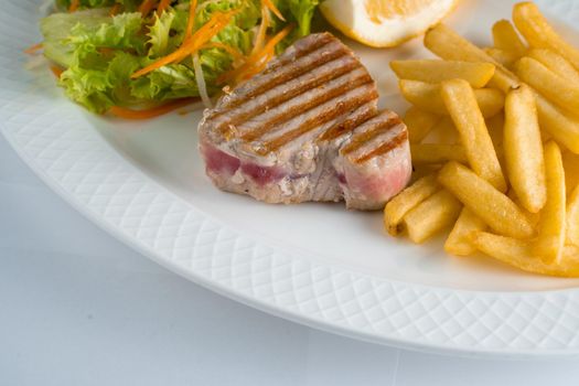 Tuna steak grilled with french fries salad of carrots, parsnips, lettuce and lemon on white plate on white background