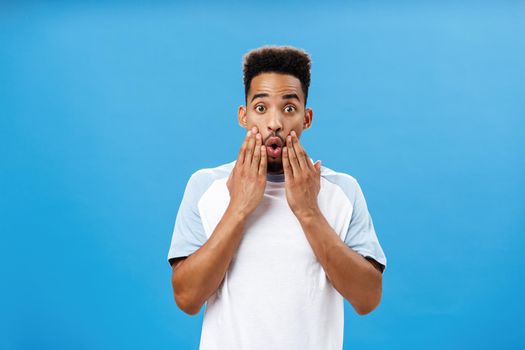 Portrait Lifestyle. Attractive young dark-skinned guy likes hearing fresh rumors learning new hot details gasping opening mouth holding hands on face staring impressed and astonished at camera over blue background.