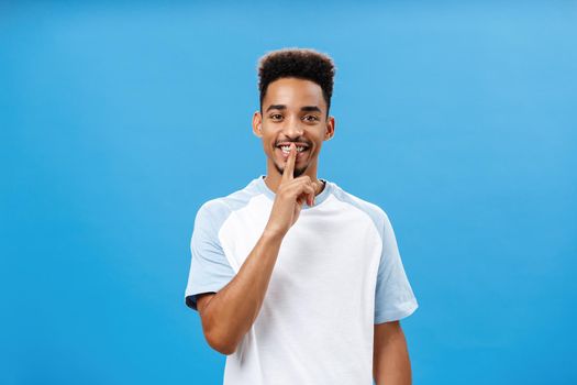 Mysterious romantic, handsome african american with afro hairstyle smiling broadly saying shh asking friend keep secret from girlfriend, make surprise making shush gesture with index finger over mouth.