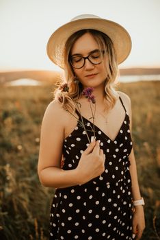 A fair-skinned blonde girl in a dress with polka dots in a hat walks through the meadow among the tall grass in the summer at sunset.