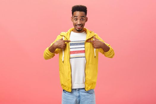 Handsome young guy being surprised he was picked. Portrait of unsure pleased charming dark skinned male with bear din stylish outfit pointing at chest with questioned expression over pink wall.