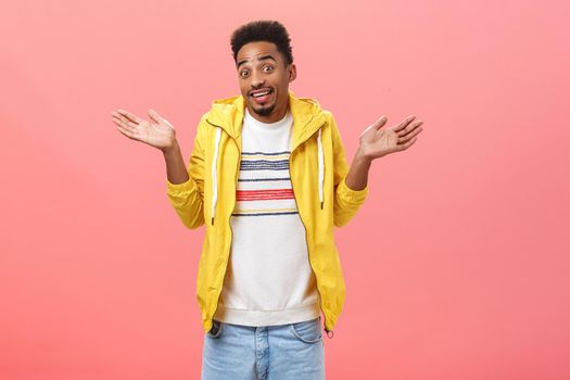 Have no idea what you talking about. Portrait of carefree unaware and clueless charming african american guy in yellow trendy jacket making shoulder shrug with spread hands being unsure over pink wall.