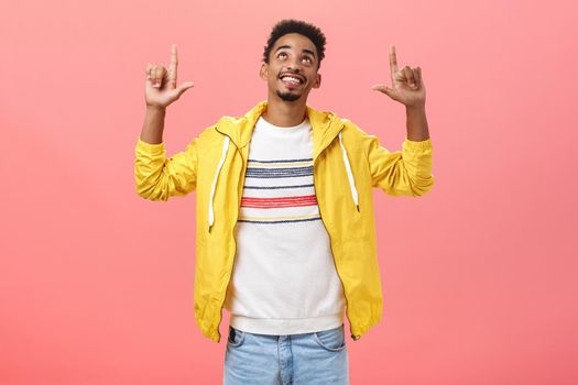 Studio shot of happy enthusiastic handsome dark-skinned male student with beard and curly hairstyle looking and pointing up with satisfied joyful expression standing over pink background. Emotions and advertising concept