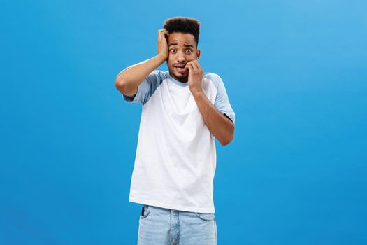 Insecure anxious guy getting scared of consequences. Portrait of frightened panicking handsome young african american male biting fingernails gazing worried at camera posing over blue background. Emotions concept