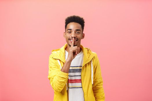 Shh do not say word. Stylish intrigued and happy good-looking african american male with beard and afro haircut smiling joyfully holding index finger over mouth in shush gesture near pink wall.