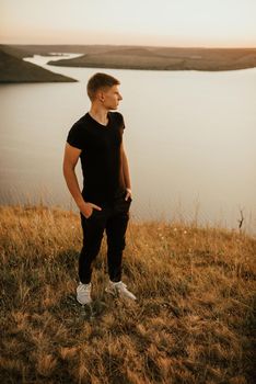 Bakota, Ukraine - 08.12.2020: A young athletic man in black casual clothes stands on the side of a mountain against the backdrop of a large lake and islands at sunset.