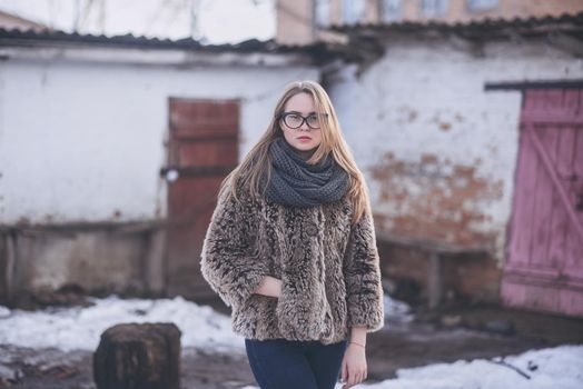 Girl blonde in cat eyes glasses in an artificial faux fur coat posing. Lying snow on the background. Red orange pink tones in the photo. Around the old shabby abandoned buildings. Cold winter