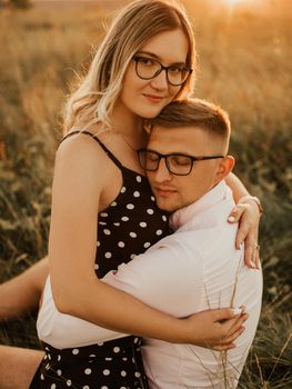 A girl in hat sits in hands guy on ground in middle of tall grass in meadow. couple of fair-haired fair-skinned people in love are resting in nature in field at sunset.