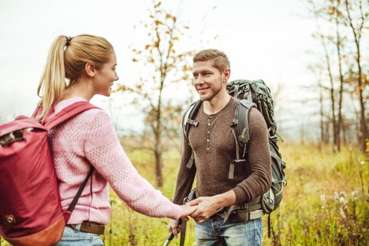 Romantic date for couple of backpackers in wild nature. Happy man and women looks each other holding hands while standing against background of autumn field. Hiking concept. Adventure concept.