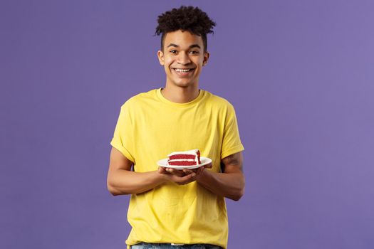 Celebration, party and holidays concept. Portrait of handsome young smiling man, feeling happy likes eating desserts, ordered delivery from local cafe to have some delicious cake, purple background.