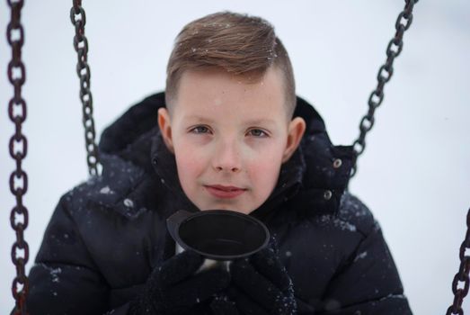 Close-up portrait of a boy holding a metal mug with a hot beverage against a background of snow.