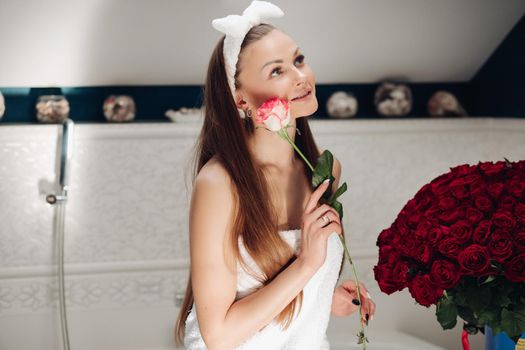 Brunette girl with long hair sitting on bath after morning shower. Pretty lady in white towel looking up and holding flower. Young woman getting bouquet of roses and thinking about boyfriend.