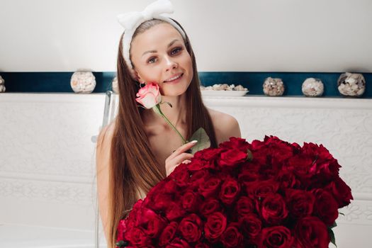 Portrait of gorgeous sensual young brunette woman with headband wrapped in towel sitting in bathroom with bunch of beautiful red roses. She is holding fragile pink rose and smiling at camera. Beauty and skincare concept.