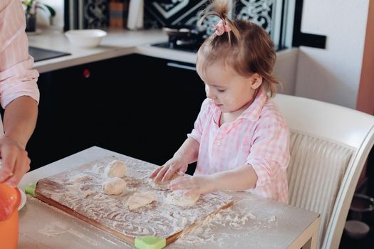 Portrait of lovable child with tail in pink checkered shirt helping mother cooking dumplings or piers in the kitchen. Sitting on chair and kneading dough on board with flour.