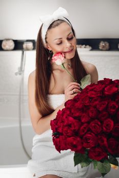 Portrait of gorgeous sensual young brunette woman with headband wrapped in towel sitting in bathroom with bunch of beautiful red roses. She is holding fragile pink rose and smiling at camera. Beauty and skincare concept.