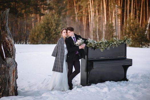 A girl in a wedding dress and a young man are standing next to the piano. Winter photography in the forest.