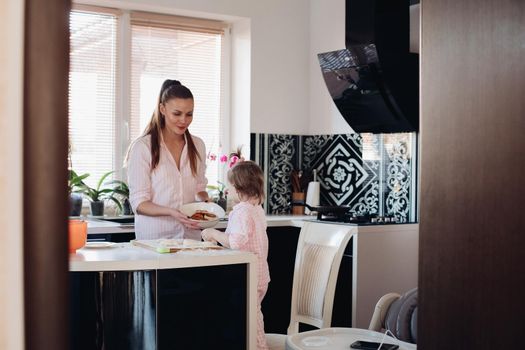 Lovely mother playing with little daughter in kitchen. Cheerful woman and cute girl cooking and baking donuts at kitchen. Happy family laughing and talking together at home. Concept of love and joy.