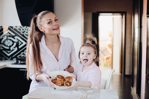 Front view of attractive mother and little daughter looking at camera and showing baked curd fritter. Happy family cooking and playing together in kitchen. Concept of love and happiness.