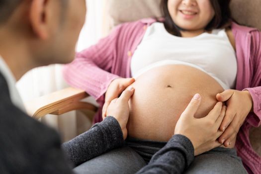 Pregnant couple of husband and wife feels love and relax at home. Young expecting woman holds baby in pregnant belly. Father take care of pregnant mother. Concept of maternity and pregnancy care.