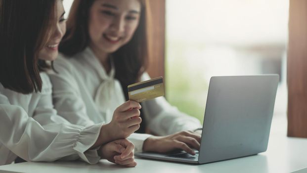Two young asian women holding credit card and using laptop computer for shopping on line with happiness, business and technology concept, digital marketing, casual lifestyle.