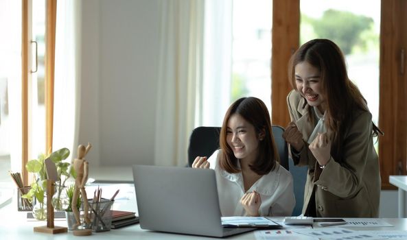 Two young Asian businesswomen show joyful expression of success at work smiling happily with a laptop computer in a modern office..