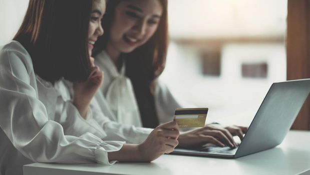 Two young asian women holding credit card and using laptop computer for shopping on line with happiness, business and technology concept, digital marketing, casual lifestyle.