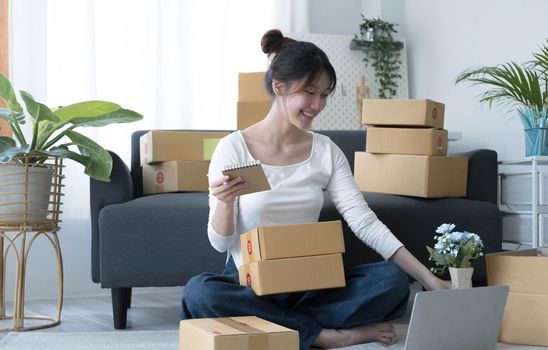 Smiling young Asian business owner woman prepare parcel box and standing check online orders of product for deliver to customer on laptop computer. Shopping Online concept..