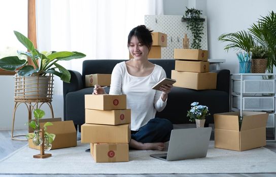 Smiling young Asian business owner woman prepare parcel box and standing check online orders of product for deliver to customer on laptop computer. Shopping Online concept..