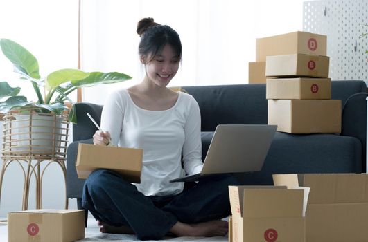 Smiling young Asian business owner woman prepare parcel box and standing check online orders of product for deliver to customer on laptop computer. Shopping Online concept..