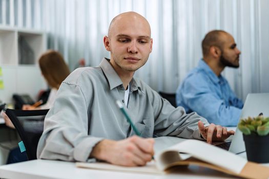 Young bald business man sitting at desk in office, working on computer, close up