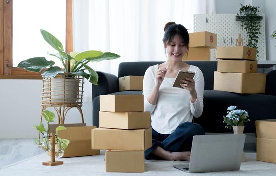 Smiling young Asian business owner woman prepare parcel box and standing check online orders of product for deliver to customer on laptop computer. Shopping Online concept..