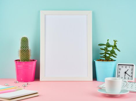 Mockup with blank white frame, alarm, notepad, cup of coffee or tea on pink table against blue wall with copy space.