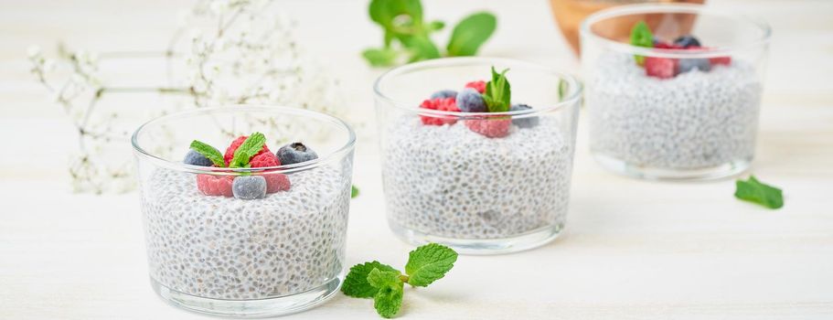 Long banner with chia pudding with fresh berries raspberries, blueberries. Three glass, light wooden background, side view, diagonal, flowers