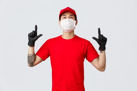 Disappointed and reluctant asian courier in red t-shirt and cap, grimacing from dislike and aversion as looking pointing up, showing unpleasant thing on top, wear uniform while deliver goods.