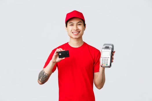 Smiling friendly courier in red uniform t-shirt and cap, advice pay for food delivery or orders with credit cards using payment terminal. Delivery guy show POS paying method, standing grey background.