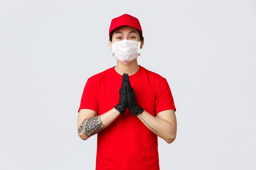 Please stay safe home, we deliver your orders. Cute asian delivery guy in red cap and t-shirt, wearing protective mask and gloves, show hands in pray, begging, pleading for something.