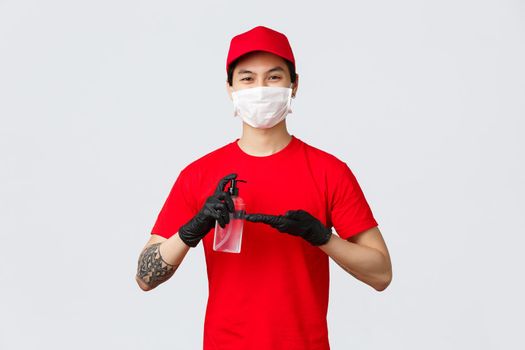 Smiling delivery man in red cap and t-shirt, wear medical mask and gloves, apply hand sanitizer to protect from covid-19, virus during outbreak. Courier cares about clients, use sanitizer during work.