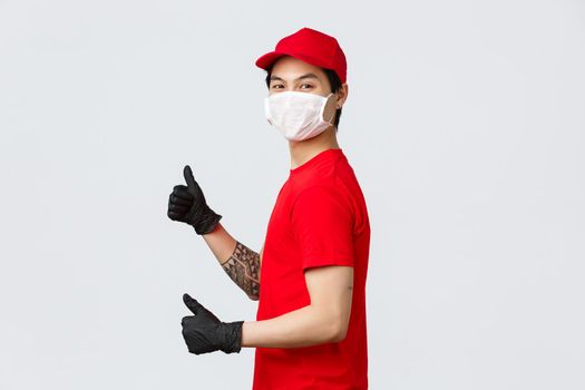 Best delivery service. Delivery guy in red cap and t-shirt, logistic company employee show thumbs-up, standing profile and turn camera with protective medical mask, gloves, recommend courier service.