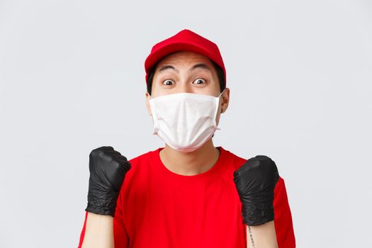 Covid-19, self-quarantine online shopping concept. Hopeful and relieved delivery guy in red cap, medical mask and gloves, fist pump achieve goal, stare amazed, received raise in courier service.