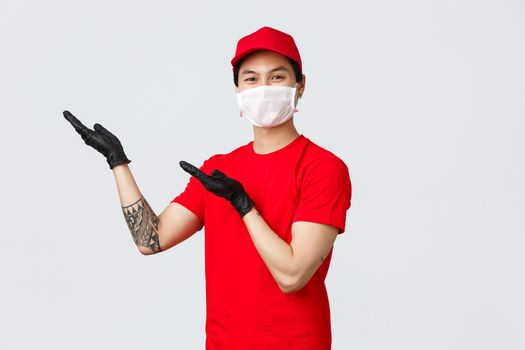 Here you are. Friendly asian delivery man in red cap, t-shirt, introduce carrier service, order tracking application, pointing hands upper left corner. Courier in medical mask and protective gloves.