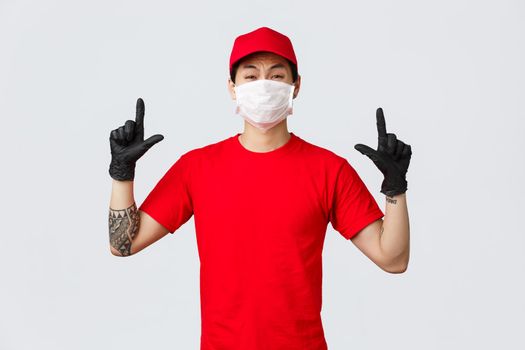 Reluctant or scared, insecure asian guy whining, grimacing as pointing fingers up, wear delivery uniform, red cap and t-shirt, company protect personnel health with protective masks and gloves.