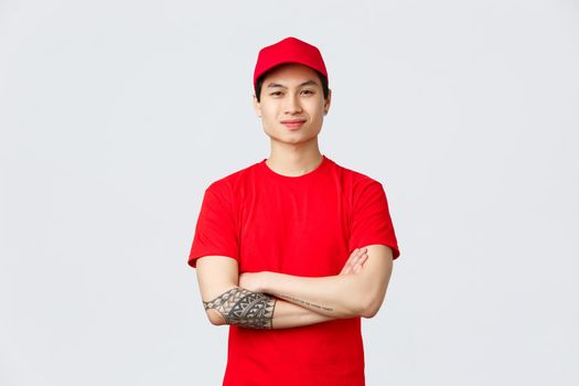Express delivery, shipping and logistics concept. Confident asian man working in deliver company, wearing red uniform with cap and t-shirt, cross arms chest and smiling.