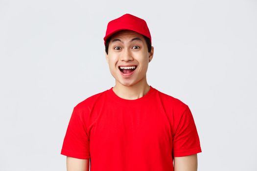 Delivery, contactless orders and shopping concept. Excited delivery guy in red uniform cap and t-shirt, courier smiling amused, look camera enthusiastic, advertise carrier service, grey background.