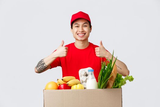 Online shopping, food delivery and internet stores concept. Smiling handsome asian courier in t-shirt, red cap, show thumbs-up in approval, guarantee quality of grocery products delivered to client.