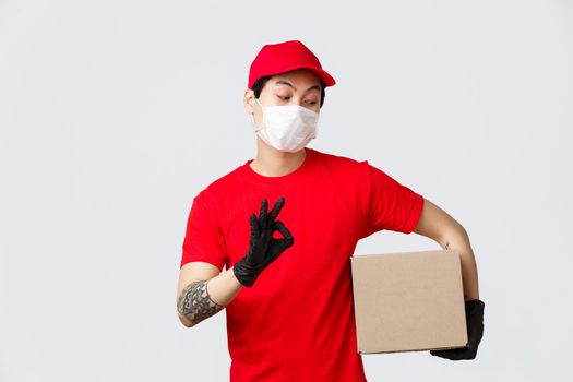 Your parcel is safe. Relaxed asian courier in medical mask and gloves, holding box package and show okay sign, guarantee best service, fast delivery, shipping online order during covid-19 outbreak.