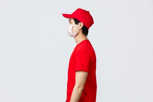 Profile shot of asian delivery guy in red cap and t-shirt, wearing protective medical mask, looking left, waiting at doorstep for client pick-up parcel, courier deliver goods during pandemic covid 19.
